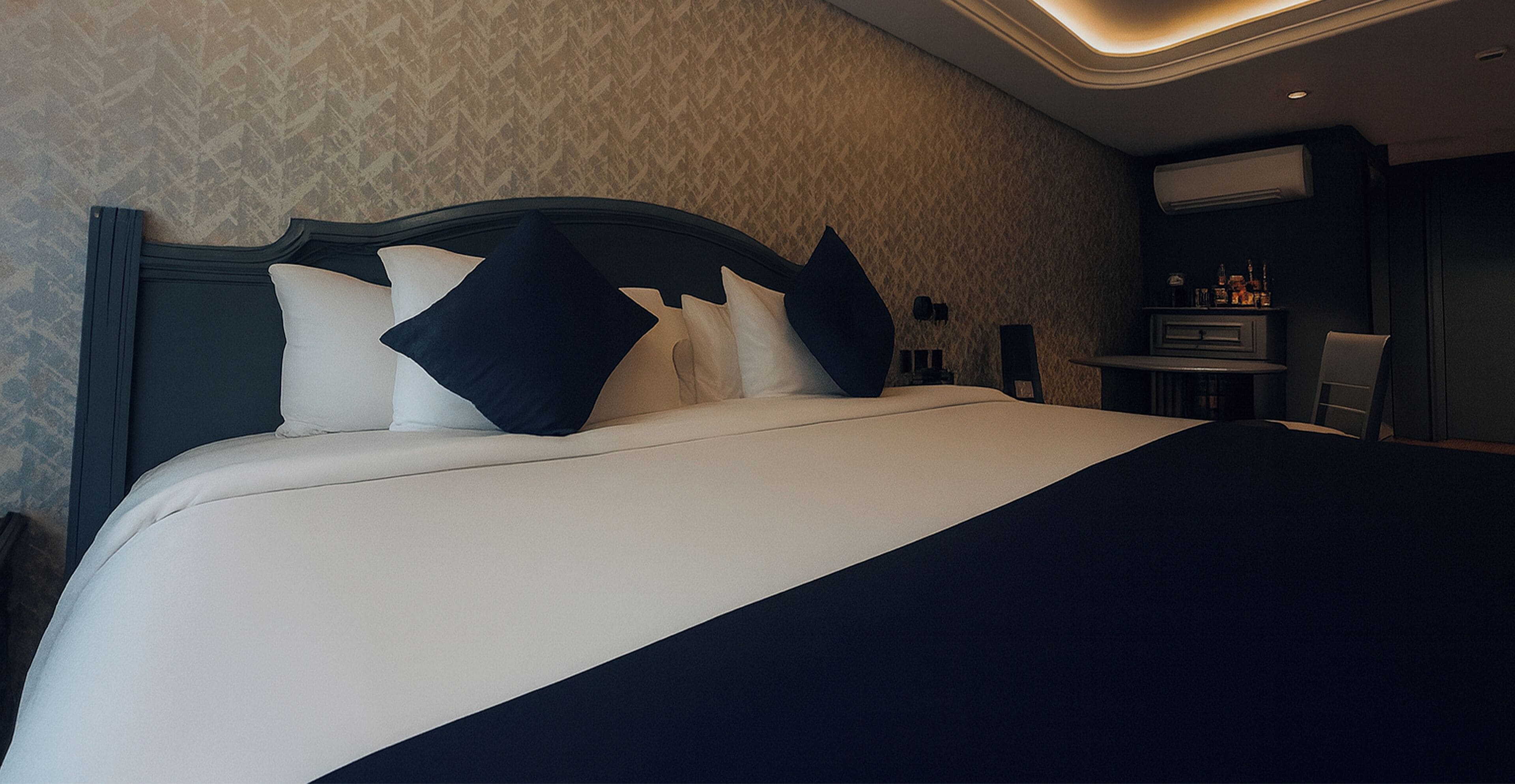 A neatly made bed with a dark headboard and white bedding in a hotel room with patterned wallpaper and a small seating area with a table and chairs.