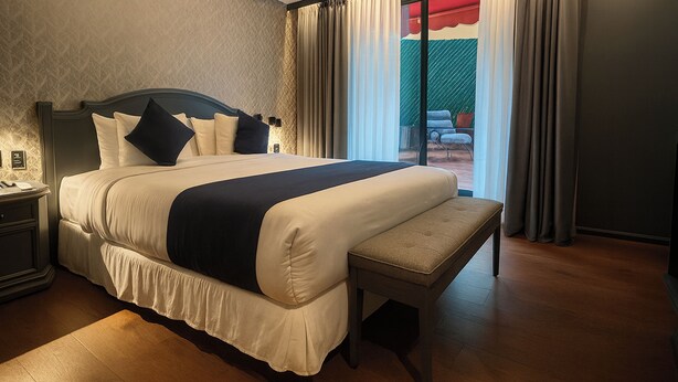 A large bed with a bench, a nightstand, and a view of the outdoors through a window. - Hive Nápoles By G Hotels (Mexico City)