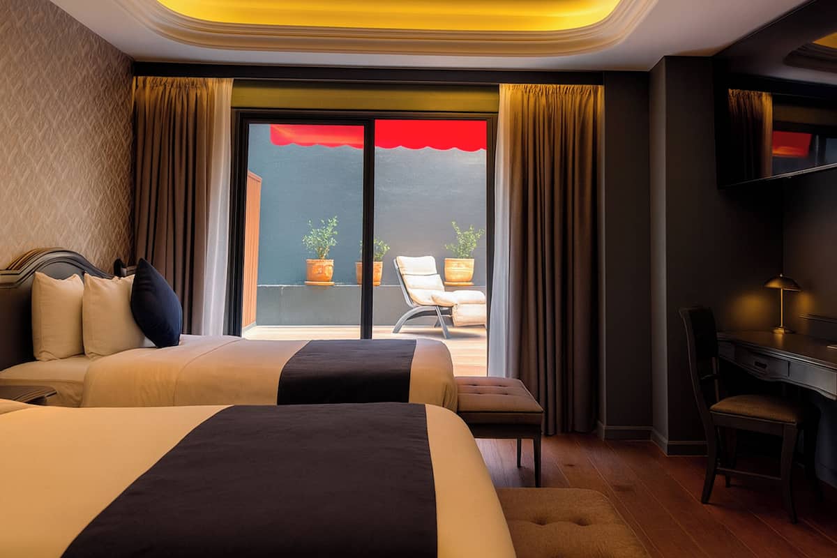 A hotel room with two beds, a desk, a chair, a TV, and a balcony with a red awning.
