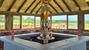 A bar area with a central island, large windows, and a view of a river and greenery. - Muthu Keekorok Lodge, Maasai Mara, Narok (Maasai Mara)