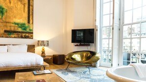 A hotel room with a bed, a flat-screen TV, a modern chair, a bathtub, and a large window with a view of the city. - Una (BRIGHTON)