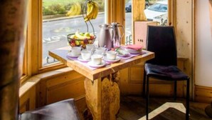 A room with a wooden table set for breakfast, a chair, and a view of buildings through a window with curtains. - Una (BRIGHTON)