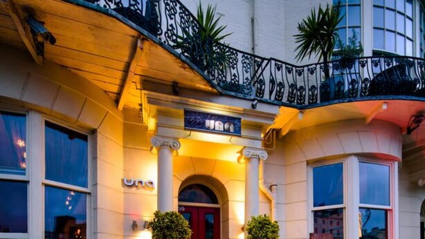 A hotel entrance with a red door, a balcony with wrought iron railings, and a sign above the door. - Una (BRIGHTON)