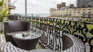 A balcony with wicker furniture and a glass table, overlooking a cityscape and the sea. - Una (BRIGHTON)