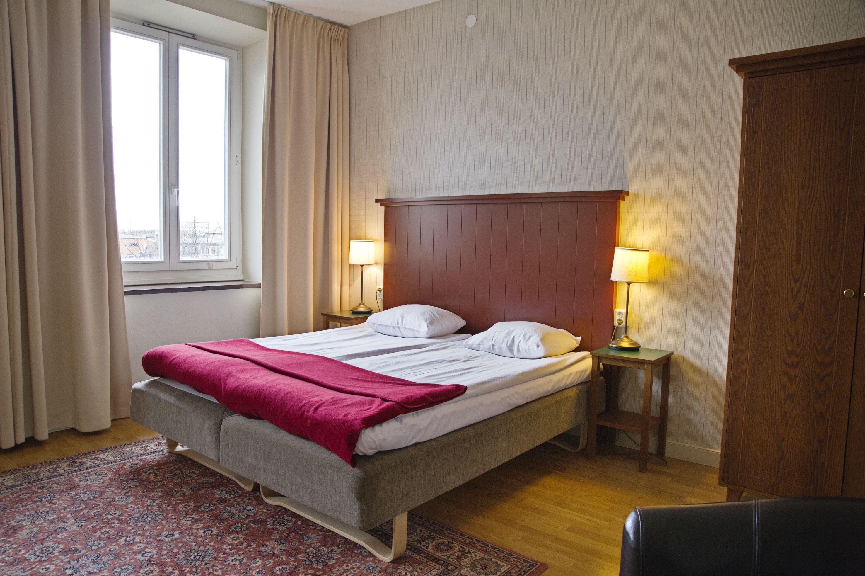 A hotel room with a bed, two bedside tables with lamps, a wooden wardrobe, a window with curtains, and a patterned rug.