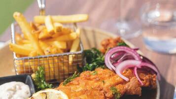 Un plato de pescado con patatas fritas, adornado con limón y cebolla roja, acompañado de un recipiente pequeño de salsa y una cesta de papas fritas.