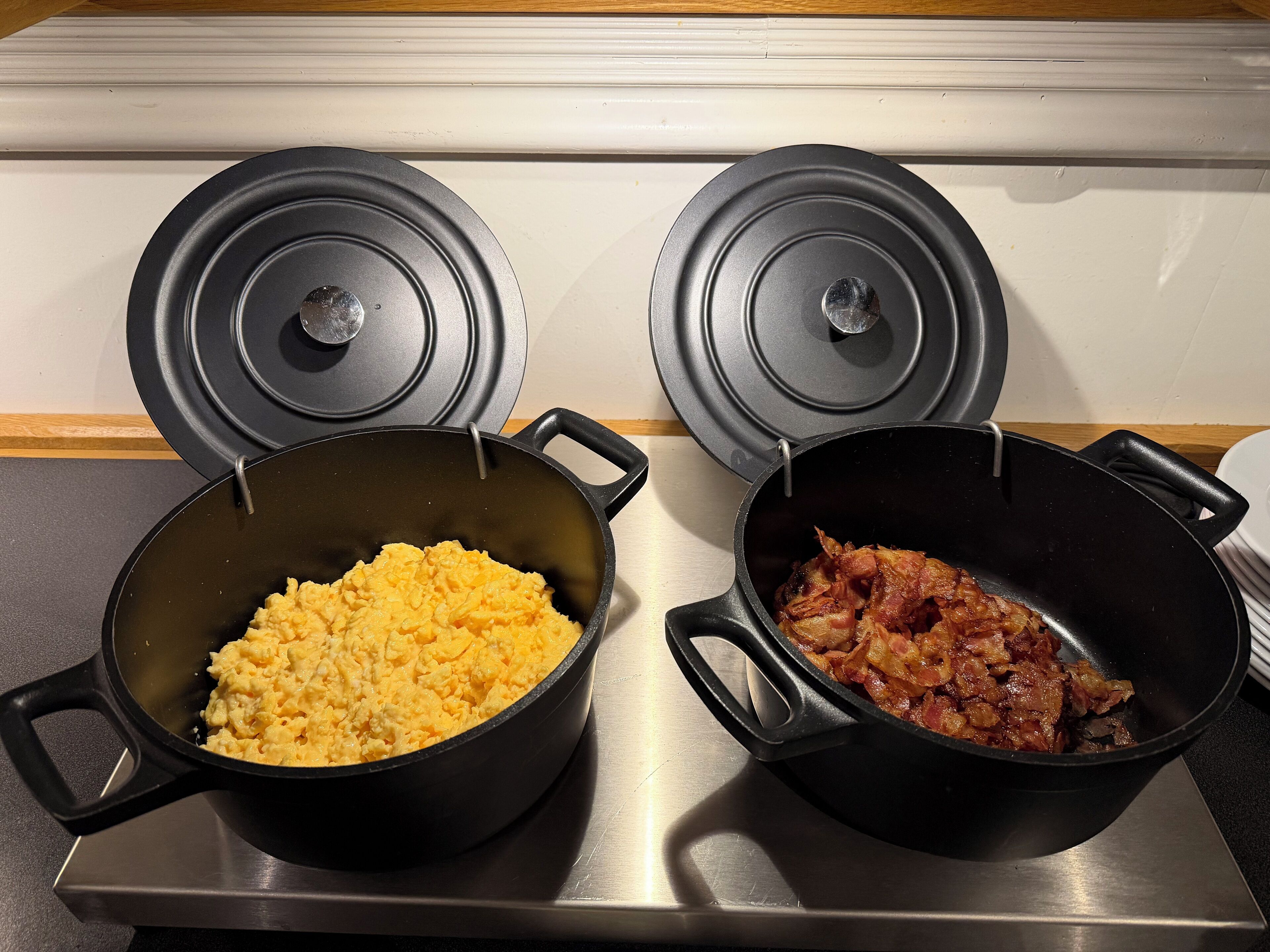 Two black pots on a stove, one with scrambled eggs and the other with bacon.