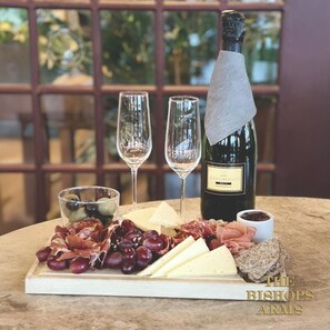 A cheese board with various cheeses, grapes, and meats, accompanied by two glasses of white wine and a bottle of wine, set on a table with a view of greenery through a window. - Hotel Bishops Arms Kristianstad (Kristianstad)
