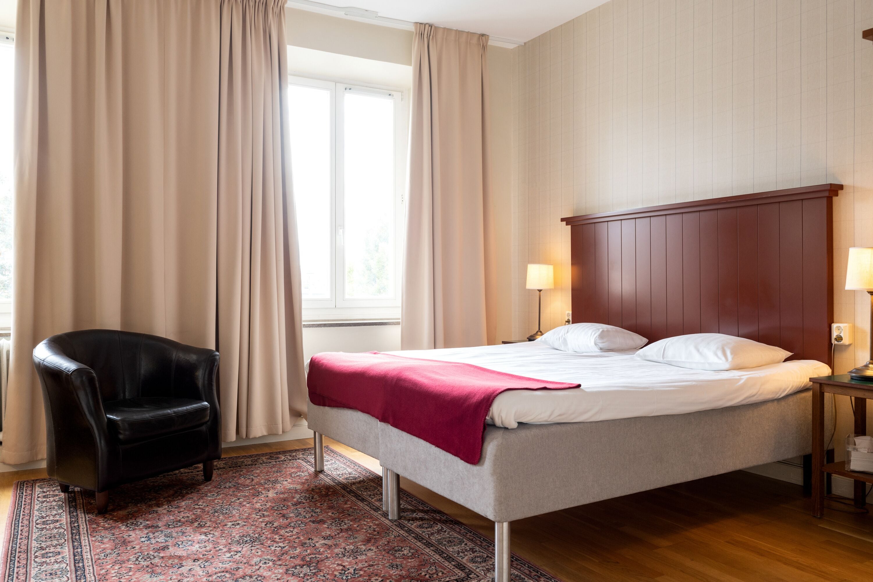 Ett hotellrum med en säng, en stol, ett nattduksbord och ett fönster med gardiner.