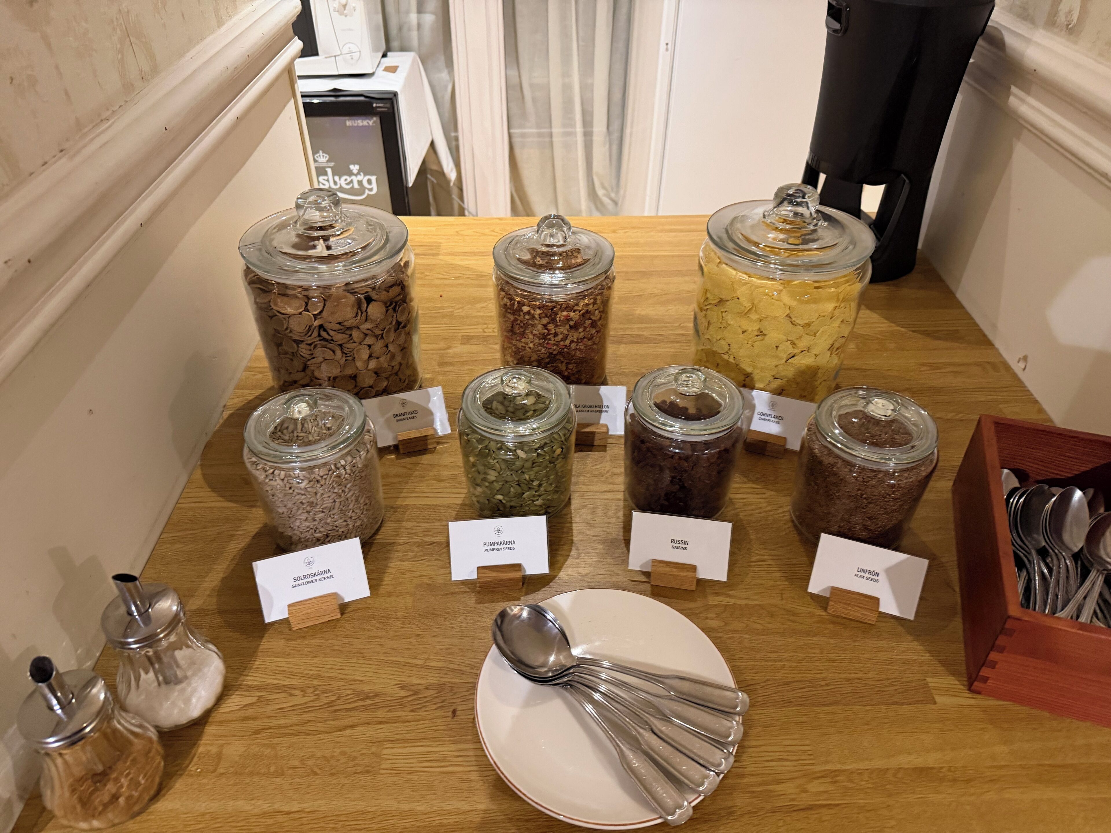 Una mesa de madera con diversos alimentos en frascos de vidrio, cada uno etiquetado con el tipo de comida y su origen.
