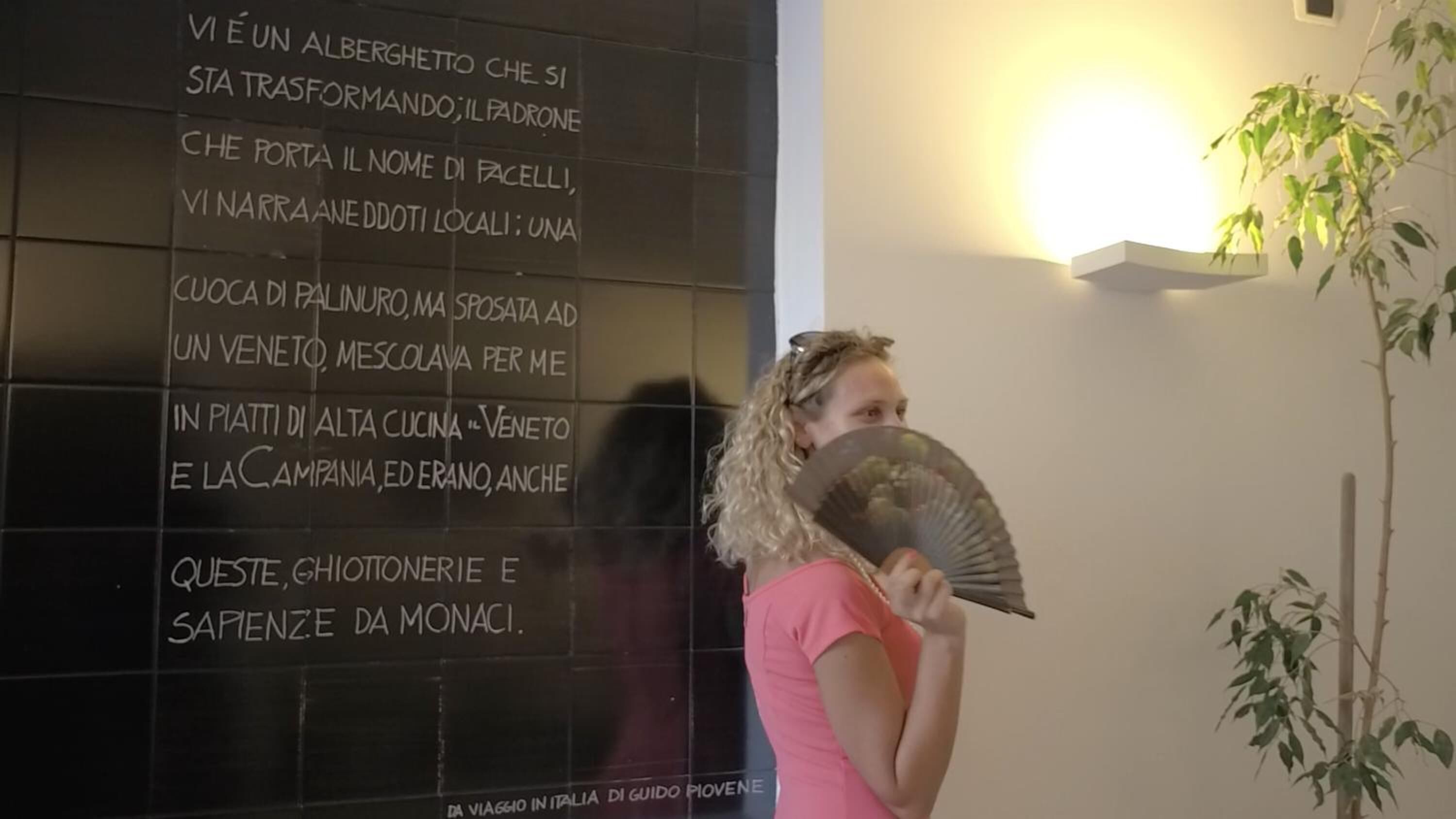 A wall with text in Italian, a person holding a fan, and a plant.