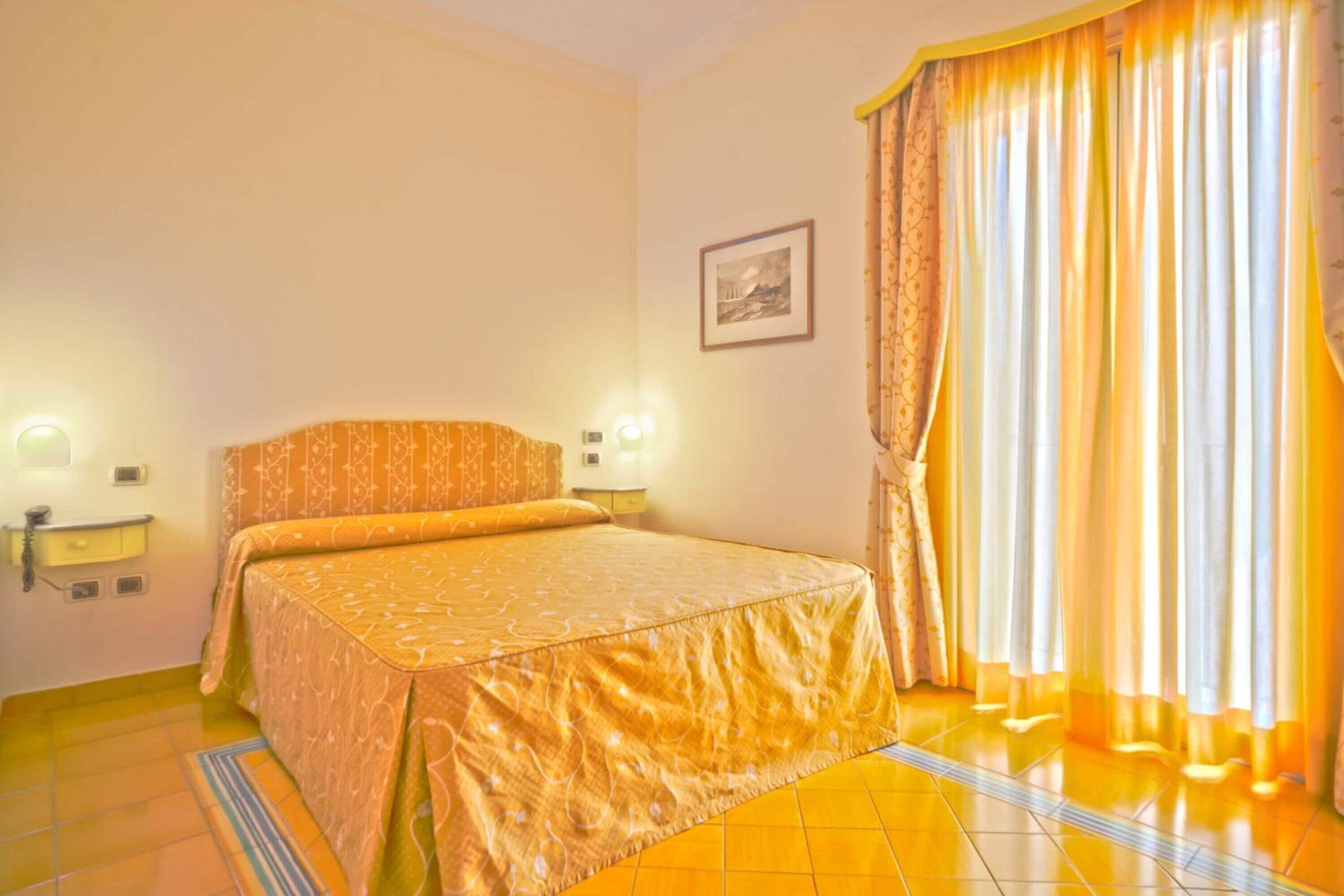 Un cuarto de hotel con una cama, dos lámparas de noche, un cuadro enmarcado en la pared y cortinas en la ventana.