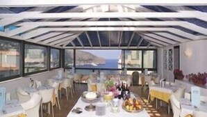 Un restaurante con vista al mar, que presenta una larga mesa dispuesta con comida y bebidas, sillas blancas y techo de cristal.