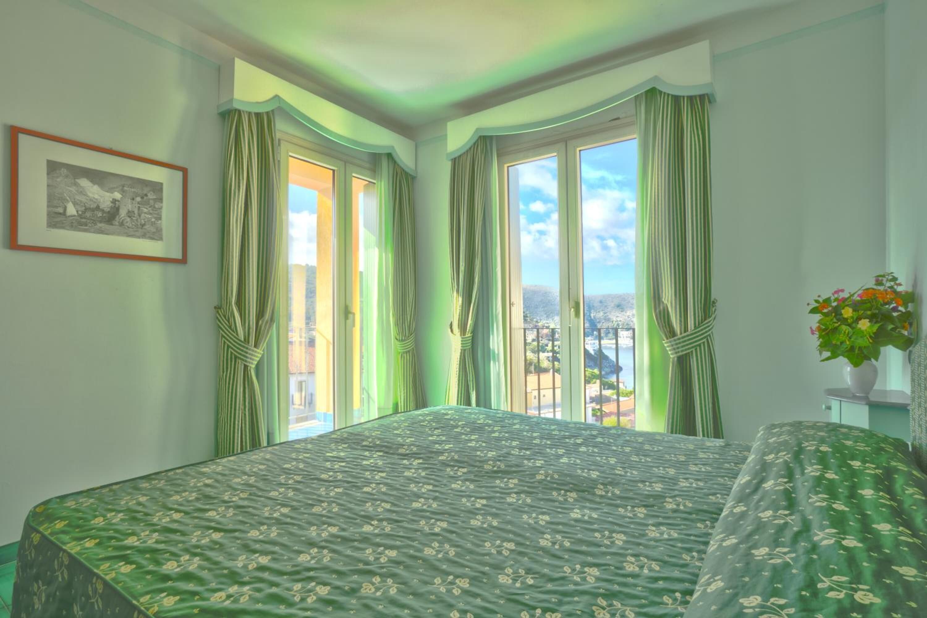 A bedroom with a large bed, two windows with curtains, a framed picture on the wall, and a potted plant on a side table.