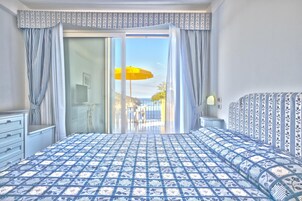 Un dormitorio con colcha estampada, una puerta corrediza de vidrio que da a un balcón con sombrilla amarilla y vista al mar.