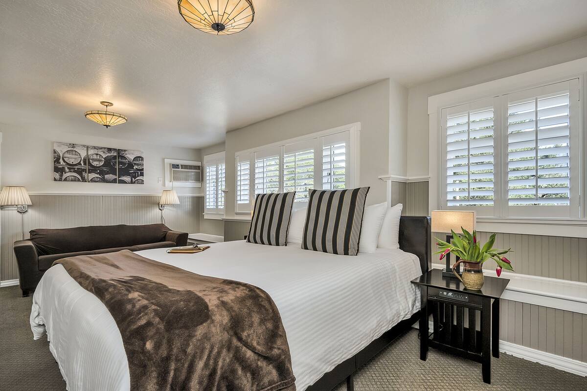A hotel room with a large bed, a bedside table with a lamp, a potted plant, and a window with blinds.