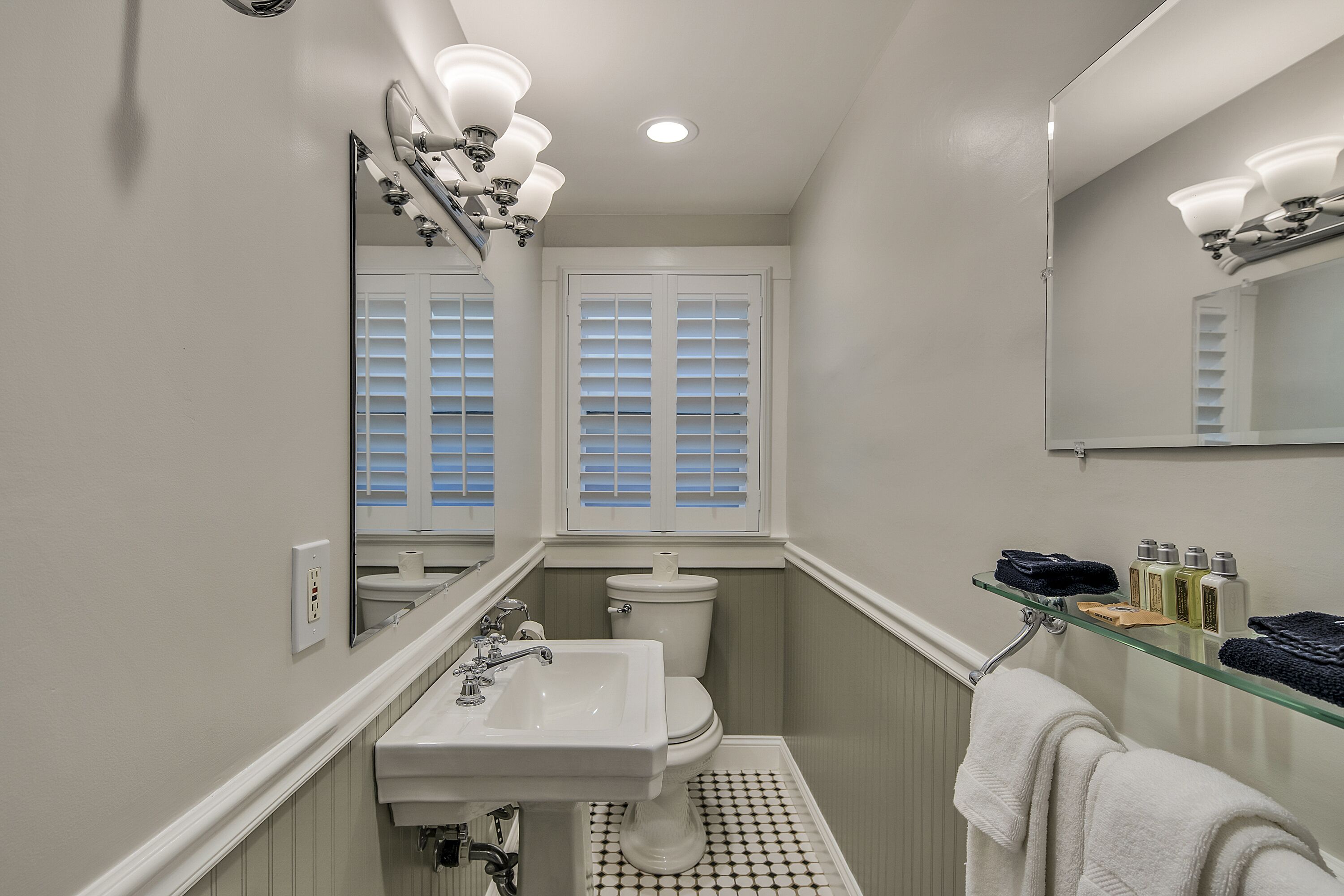 Une salle de bain avec un lavabo, des toilettes et un miroir, tous de couleur blanche. On y trouve une fenêtre avec des stores, un porte-serviettes et une étagère contenant des articles de toilette.