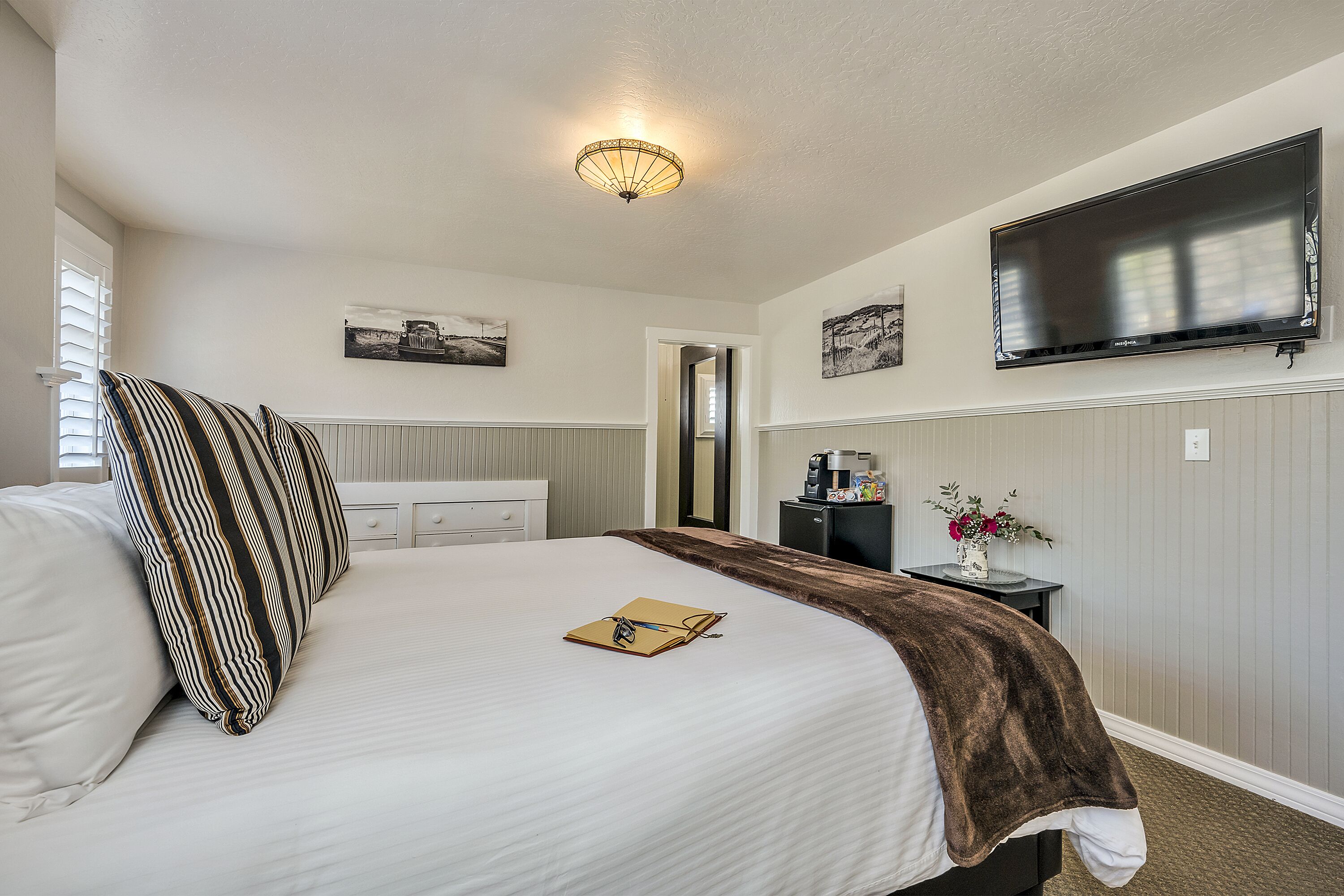 A neatly made bed with striped pillows, a brown throw blanket, and a small book and glasses on the pillow. A flat-screen TV is mounted on the wall above the bed.