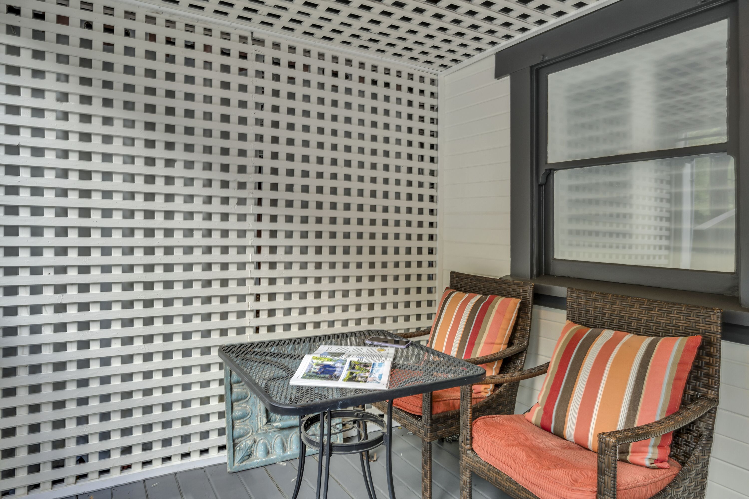 Un espacio de estar al aire libre techado con una mesa y sillas de mimbre, una pared con celosía y una ventana con vistas a edificios.