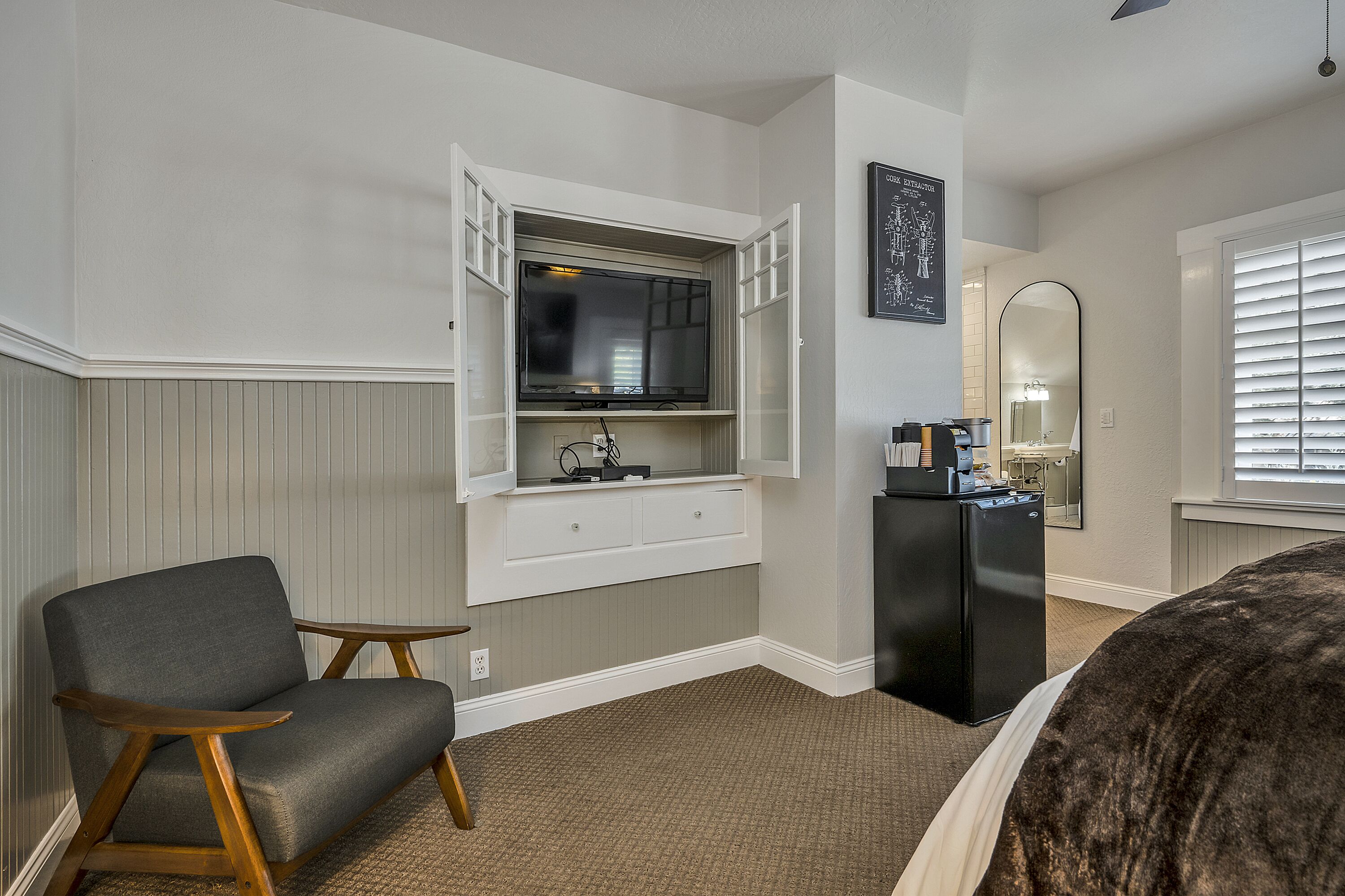 A hotel room with a built-in TV, a small desk with a refrigerator, a chair, and a bed.