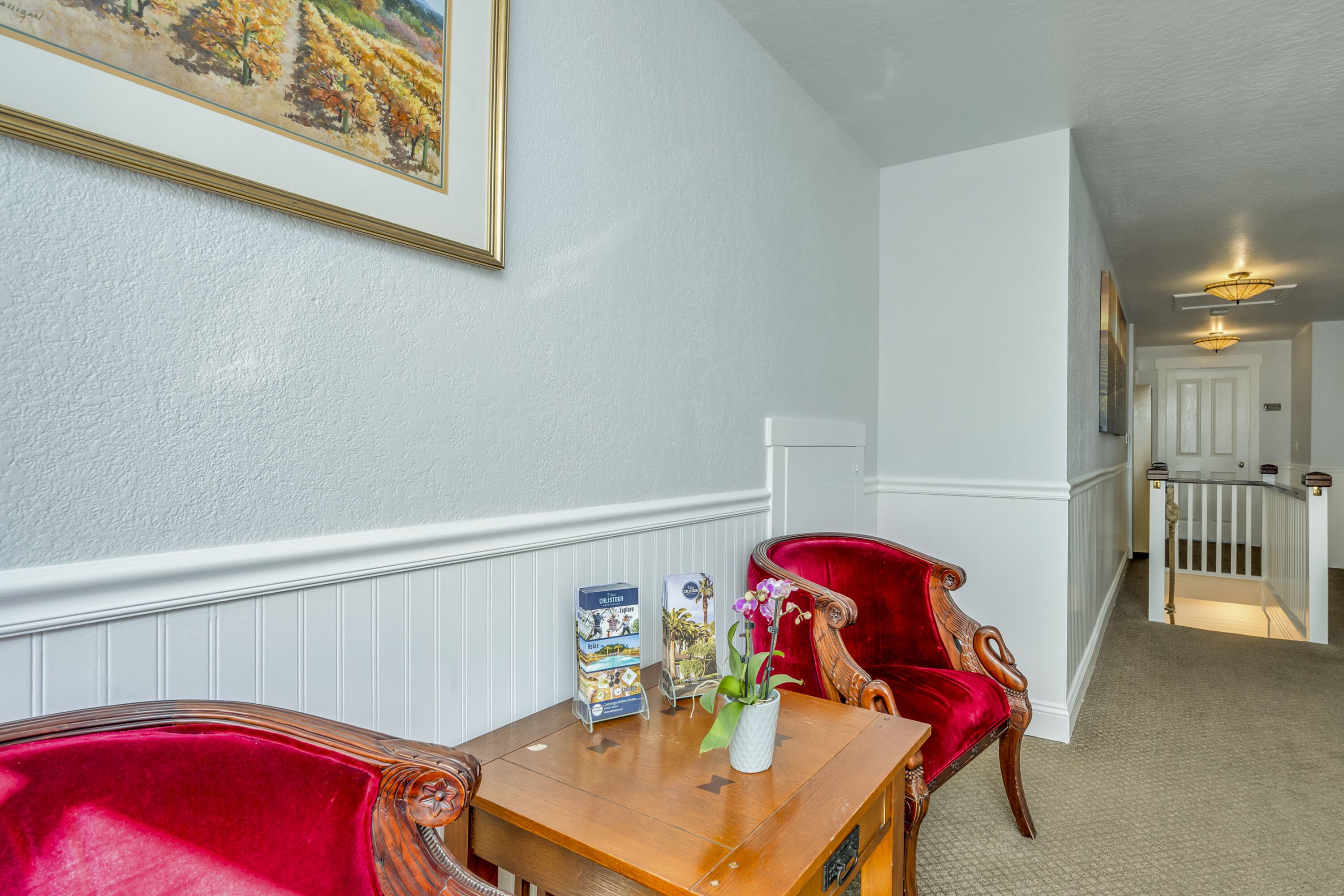 Um corredor com uma mesa de madeira, cadeiras vermelhas, um quadro emoldurado e uma escada.
