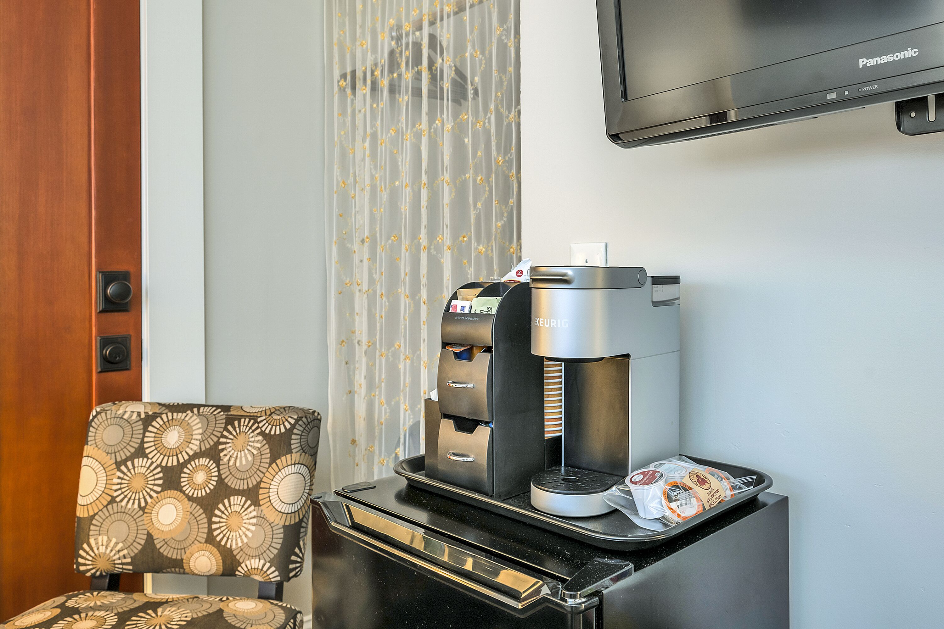 A compact kitchenette with a coffee machine, a tray with a coffee pod, and a patterned chair.