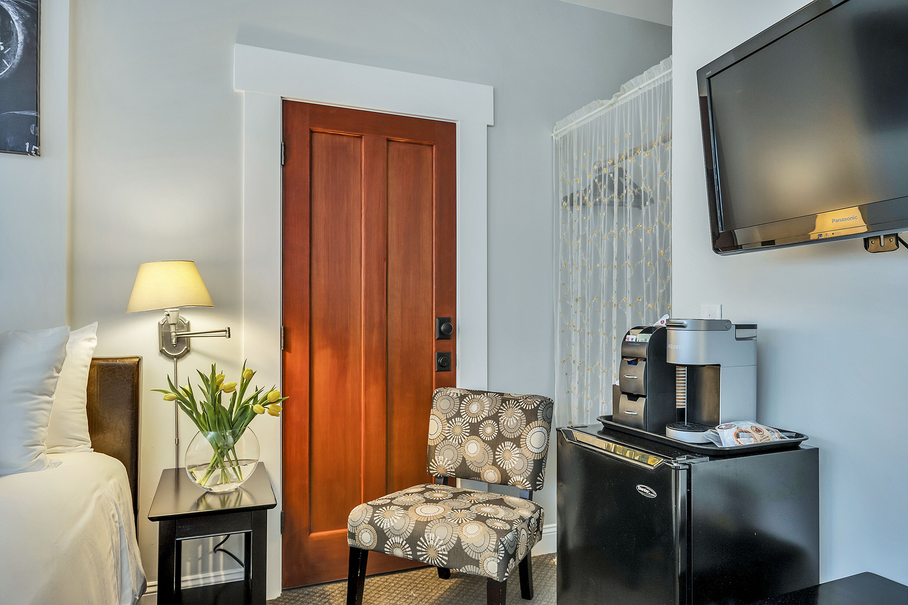 A hotel room with a bed, a desk with a lamp, a chair, a coffee machine, a TV, and a door.