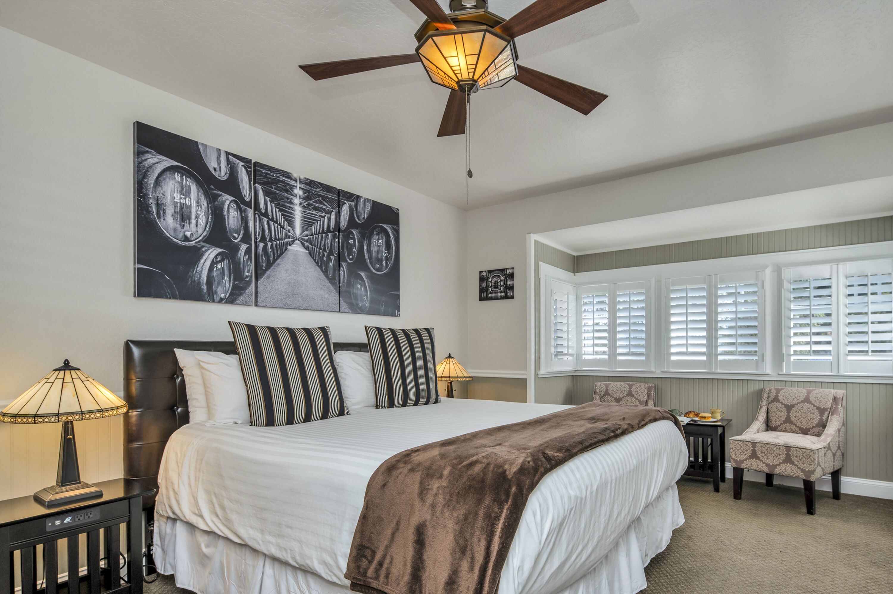 A bedroom with a large bed, a ceiling fan, a wall-mounted artwork, a window with blinds, and a chair.