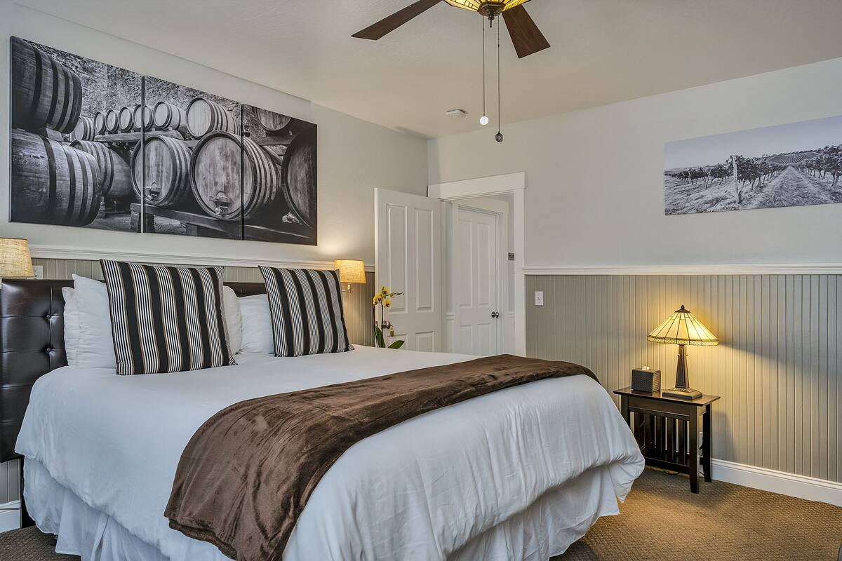 A hotel room with a large bed, a ceiling fan, a nightstand with a lamp, and two wall-mounted paintings.