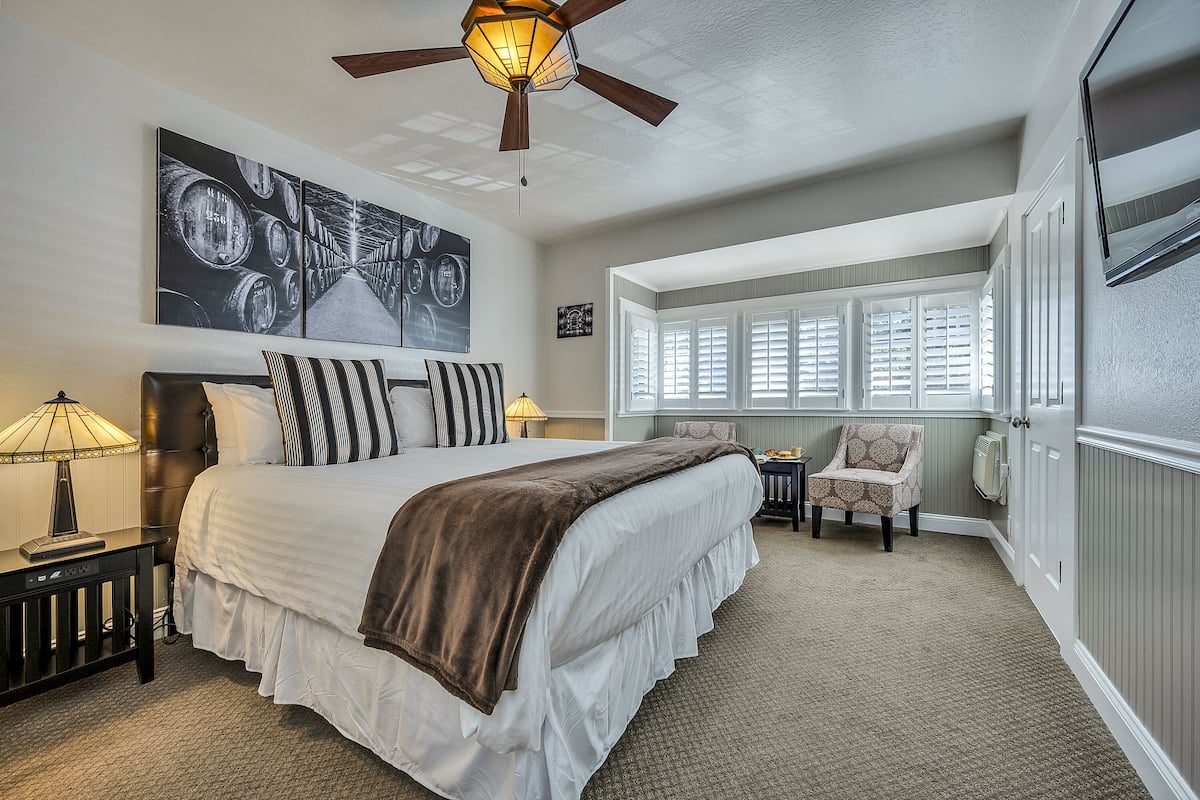 A bedroom with a large bed, a ceiling fan, a window with blinds, a chair, and a wall-mounted TV.