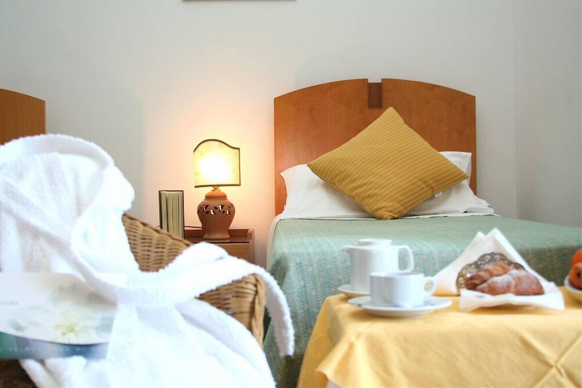 Habitación de hotel con una cama, una mesita de noche con una lámpara, una taza de café y una cesta de bollería.