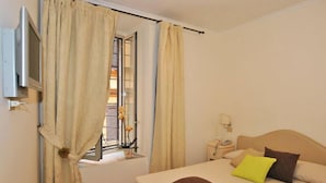 Un dormitorio con cama, cortinas, mesita de noche, lámpara y televisor.