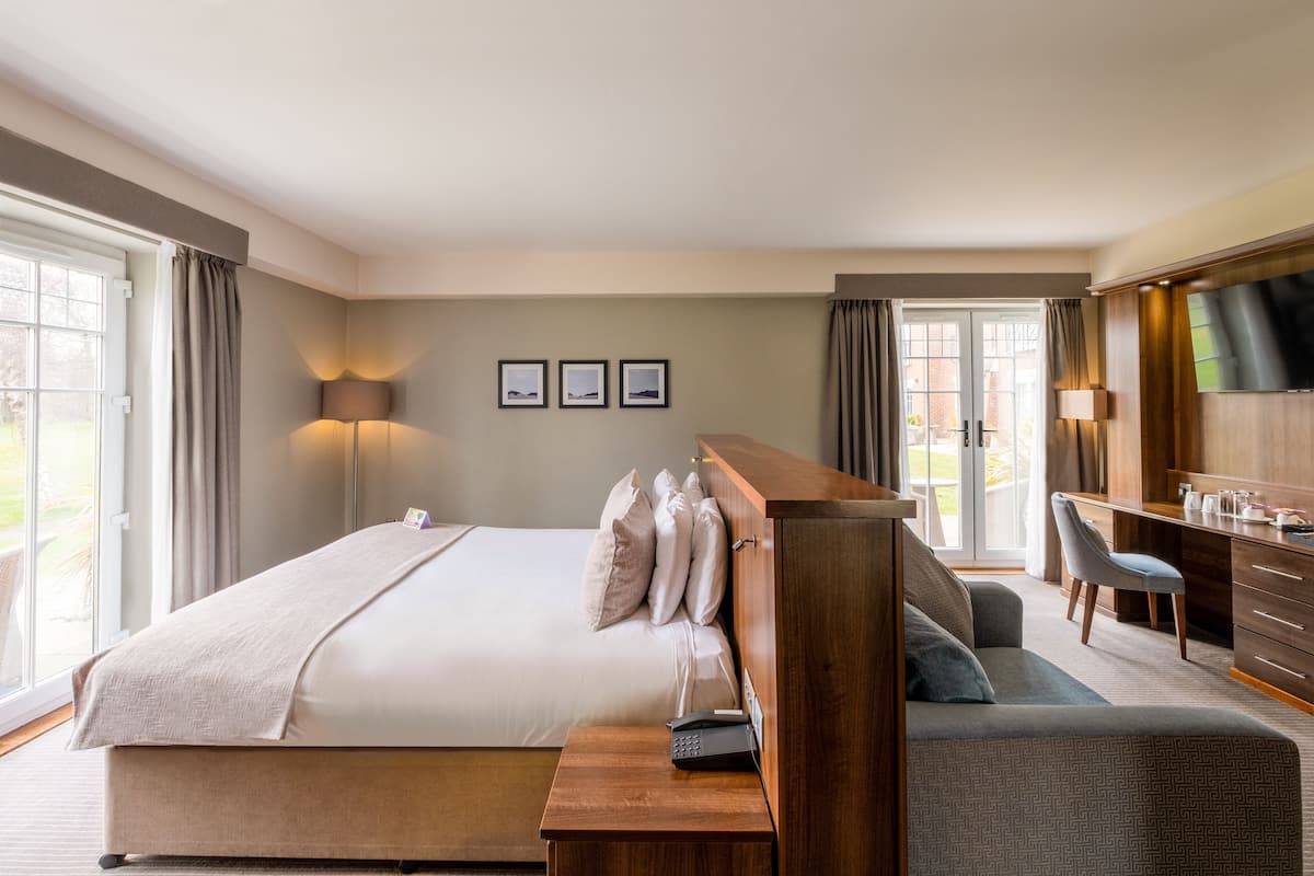 Ein modernes Hotelzimmer mit einem großen Bett, einem Schreibtisch und einem Sessel. Vorhänge, eine Lampe und gerahmte Bilder schmücken die Wand.