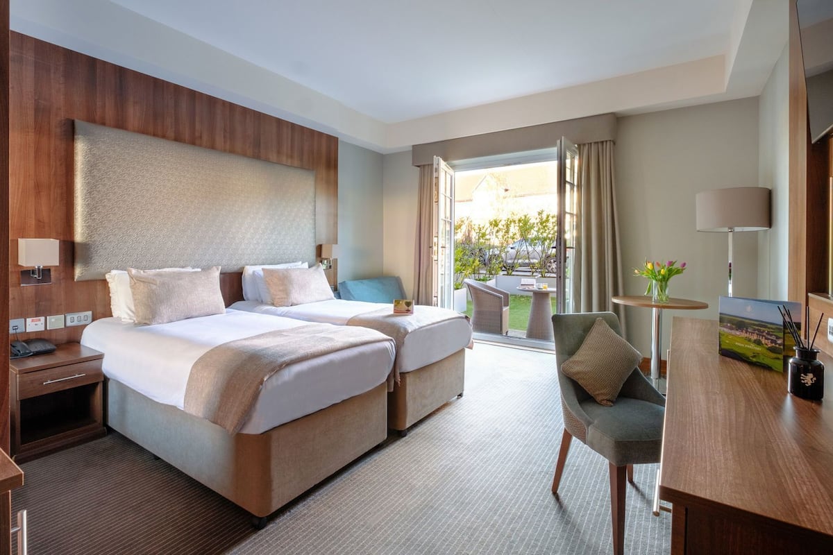 A hotel room with two beds, a desk, a chair, a lamp, and a view of the outdoors.