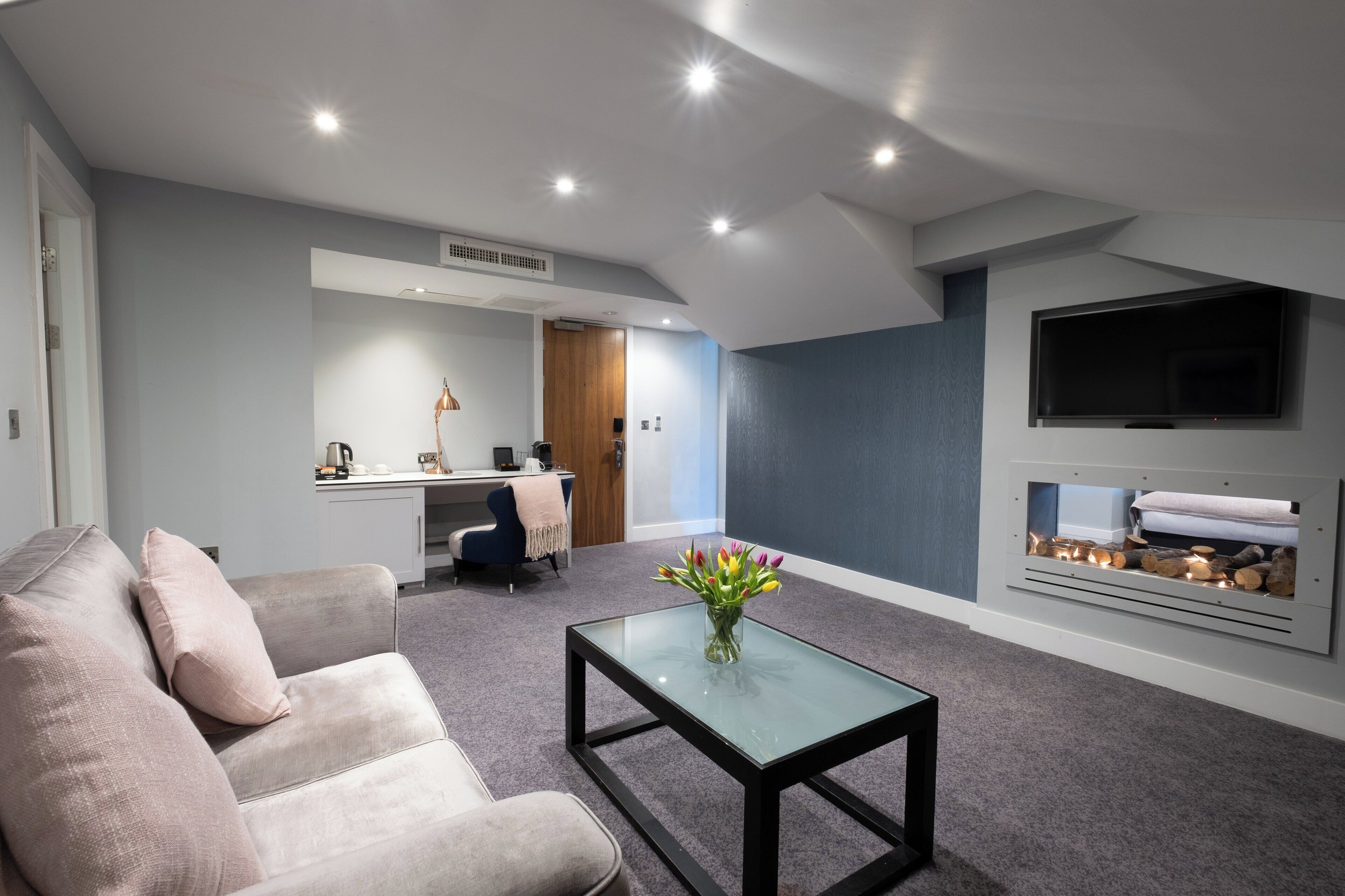 A modern living room with a sofa, a glass coffee table, a TV mounted on the wall, and a fireplace.
