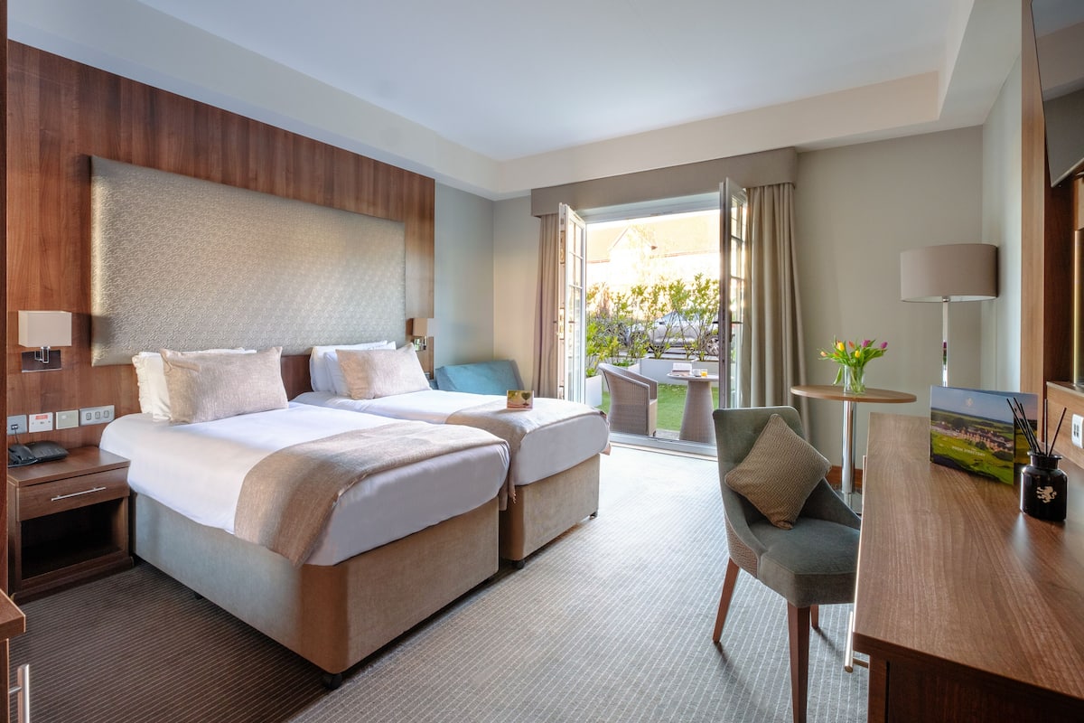 Ein Hotelzimmer mit zwei Betten, einem Schreibtisch, einem Stuhl, einer Lampe und Blick ins Freie.