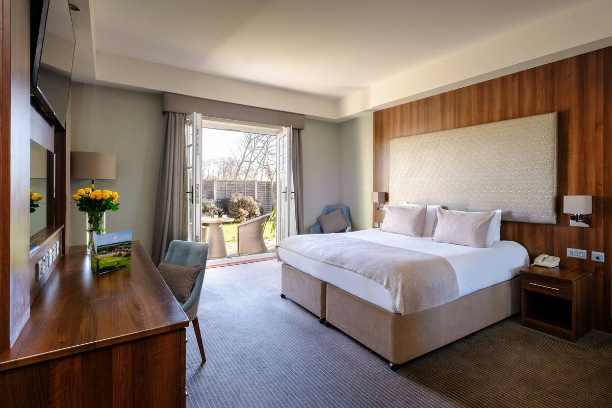 Ein Hotelzimmer mit einem großen Bett, einem Schreibtisch mit Lampe, einem Sessel, einem kleinen Tisch mit Blumen und Blick ins Freie.