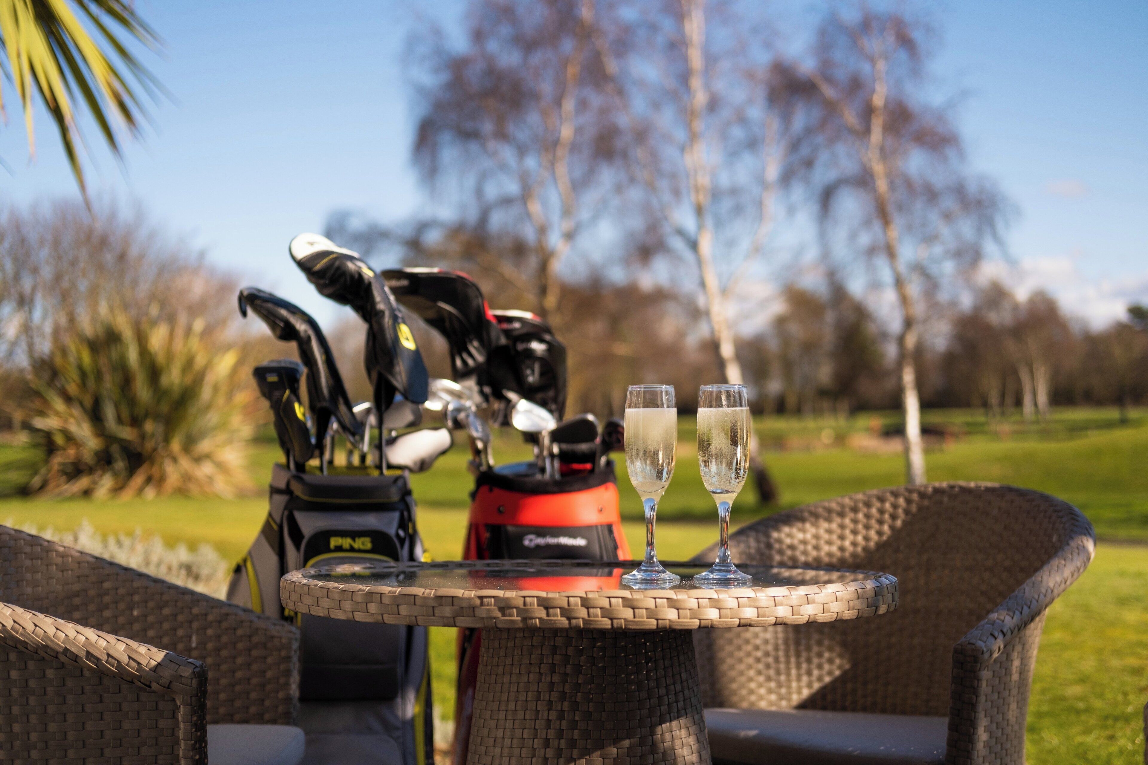 A wicker chair and table with golf clubs and two glasses of champagne on a golf course.