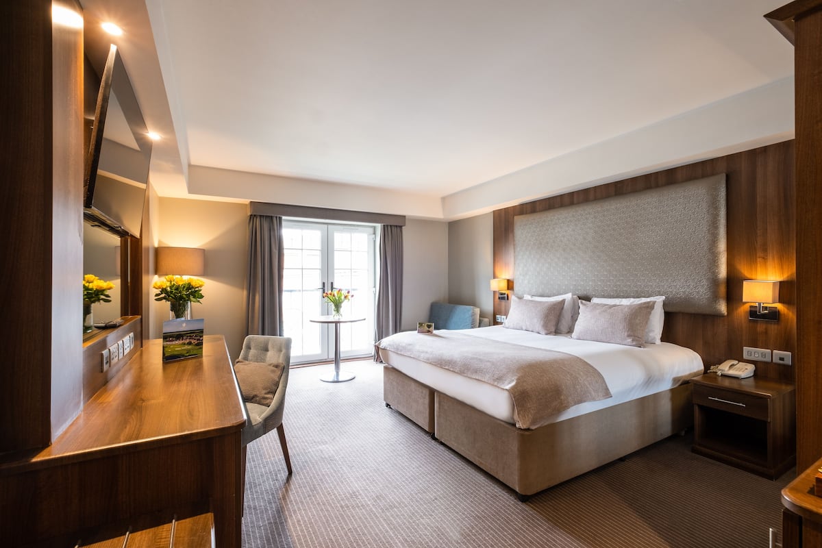 Ein modernes Hotelzimmer mit einem großen Bett, einem Schreibtisch, einem Stuhl, einem kleinen Tisch mit Blumen und einem Fenster mit Vorhängen.