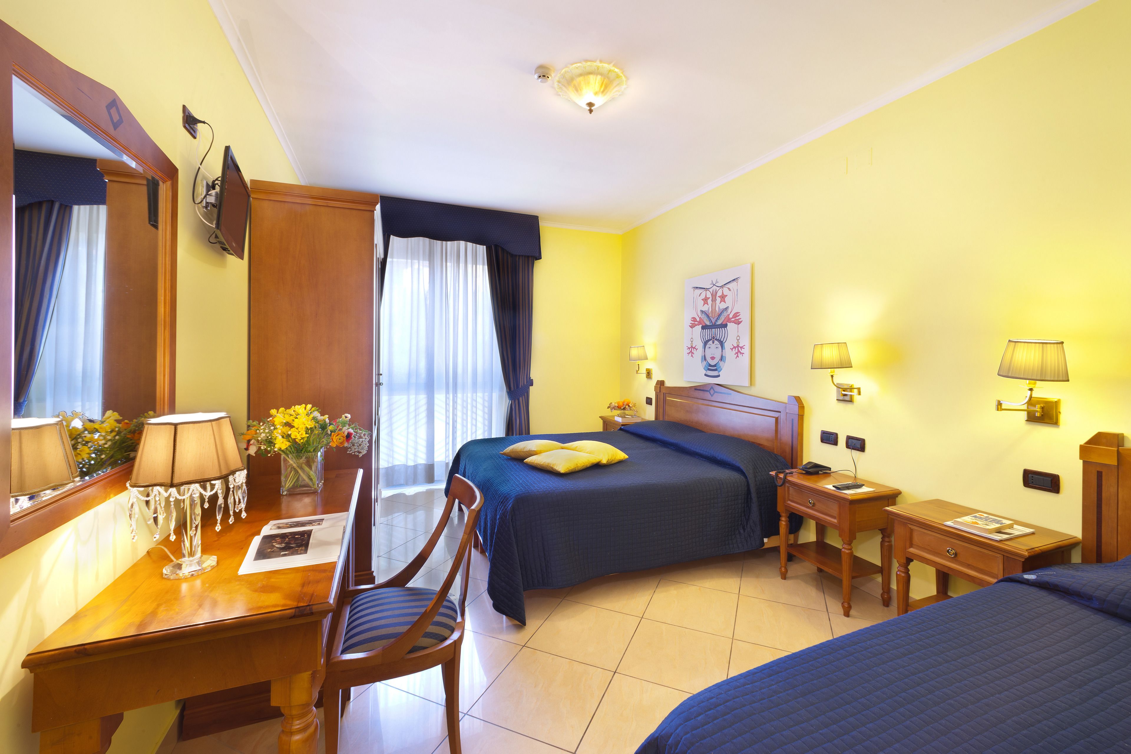 A hotel room with a bed, a desk with a lamp, a chair, a nightstand, and a painting on the wall.
