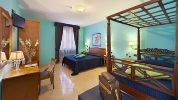 A hotel room with a four-poster bed, a desk with a lamp, a chair, and a bedside table with a vase of flowers.