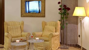 Una habitación con dos sillones, una mesita con una vela y una bebida, un florero con flores, una lámpara y un cuadro en la pared.