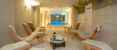 Zona de spa con sillones de descanso y una mesa central, piscina al fondo y un elemento decorativo de pared de piedra.