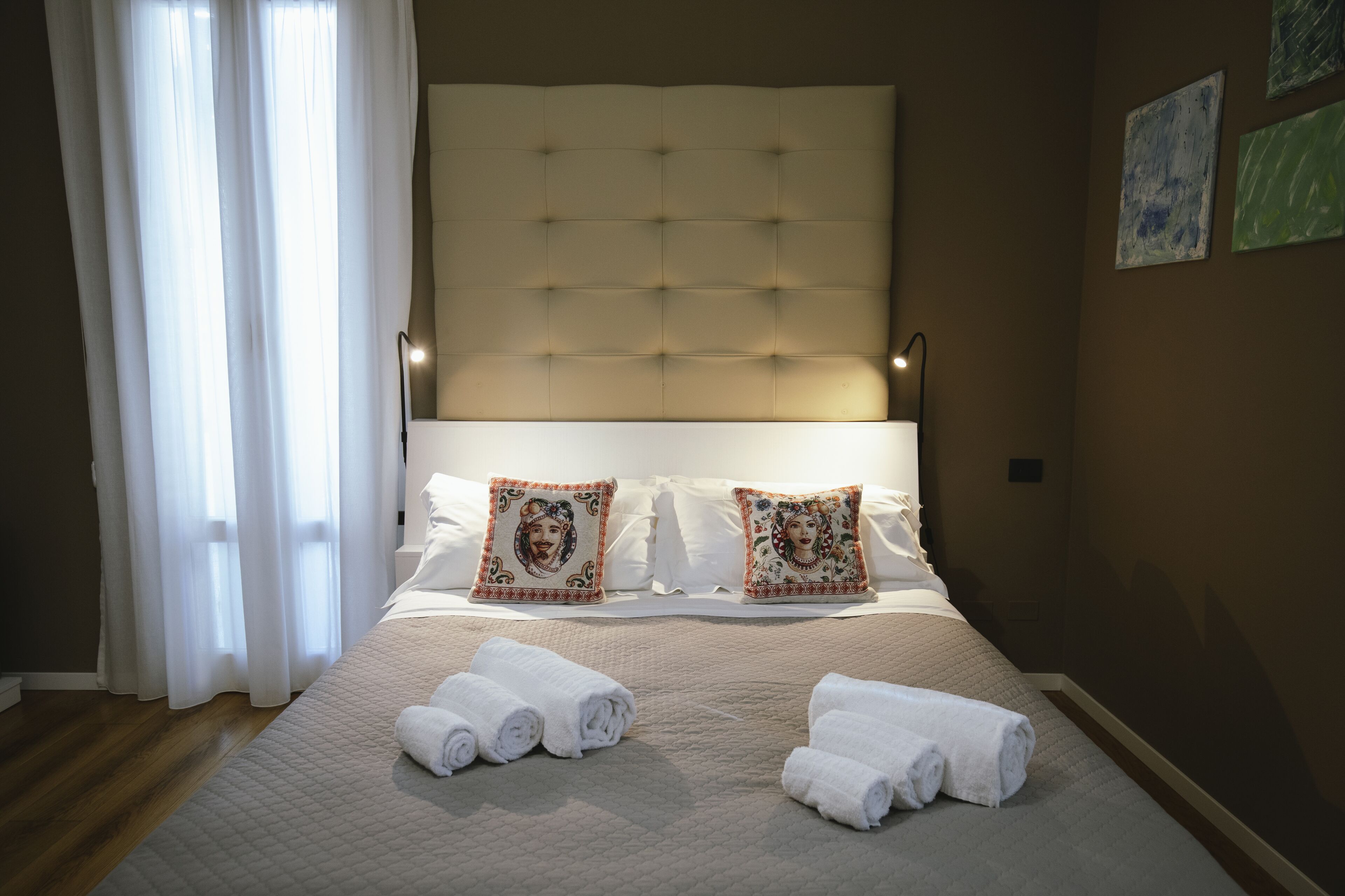 A neatly made bed with white linens and decorative pillows, a bedside lamp, and rolled towels on the floor.