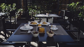 Un área de comedor al aire libre con mesas preparadas para el desayuno, incluyendo cruasanes, fruta y café.