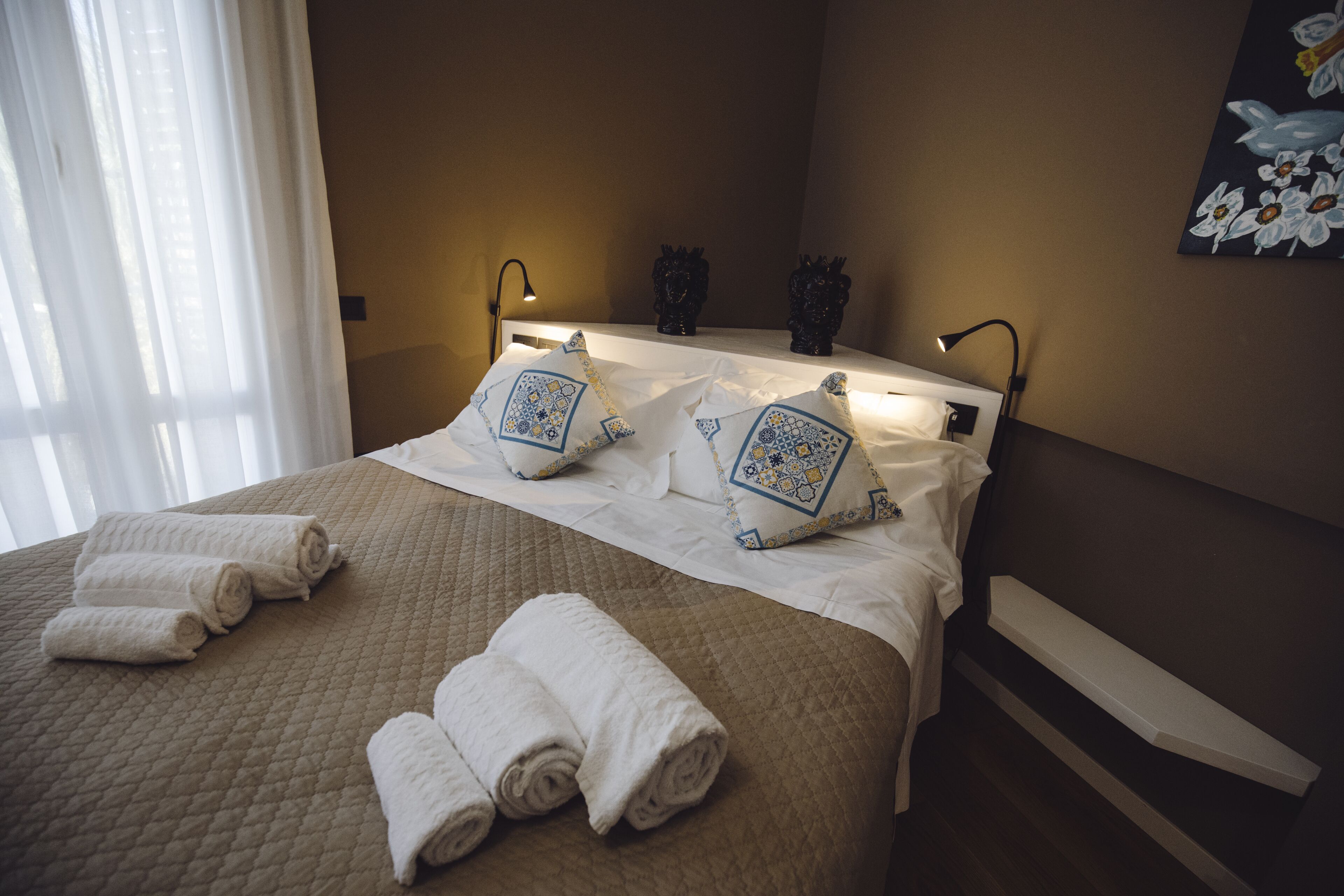 A neatly made bed with white linens and decorative pillows, a bedside table with a lamp, and a painting on the wall.