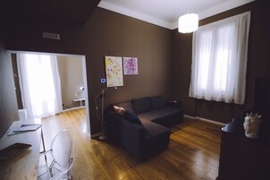 Un soggiorno moderno con un divano scuro, una sedia trasparente in vetro e un pavimento in legno. Si nota una scrivania bianca con una lampada e un piccolo tavolino con un oggetto decorativo. Due opere d'arte sono appese al muro, e una grande finestra con tende bianche è visibile.