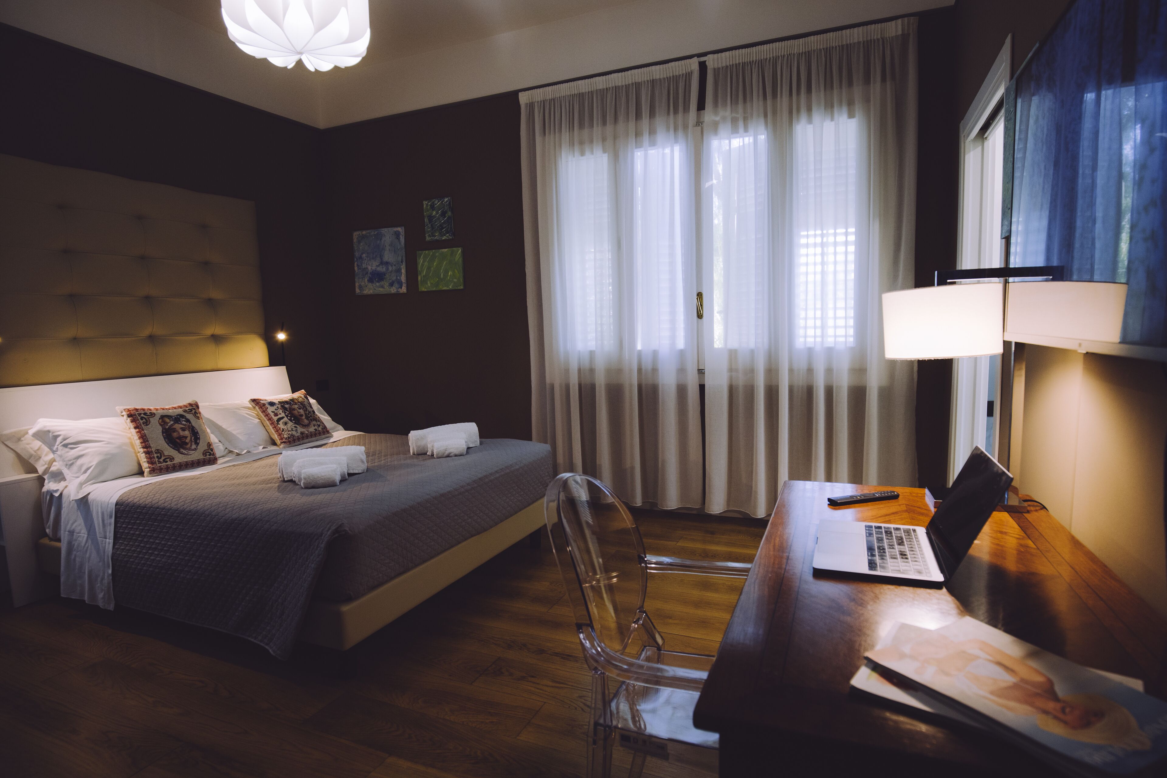 A hotel room with a bed, a desk with a laptop, a chair, a lamp, and a window with curtains.