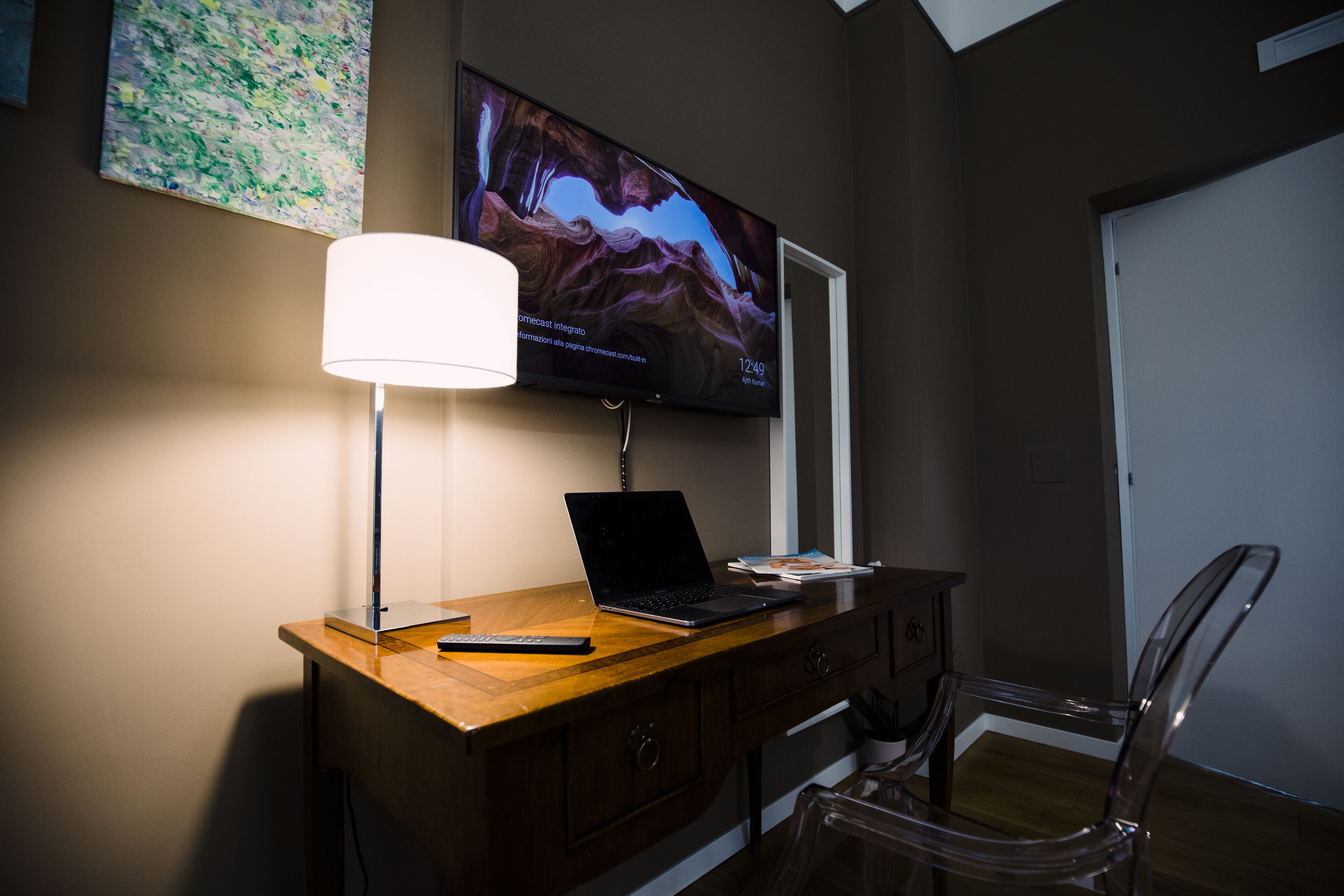 Una habitación moderna con un escritorio de madera, una computadora portátil, un control remoto y un televisor montado en la pared mostrando una imagen panorámica.
