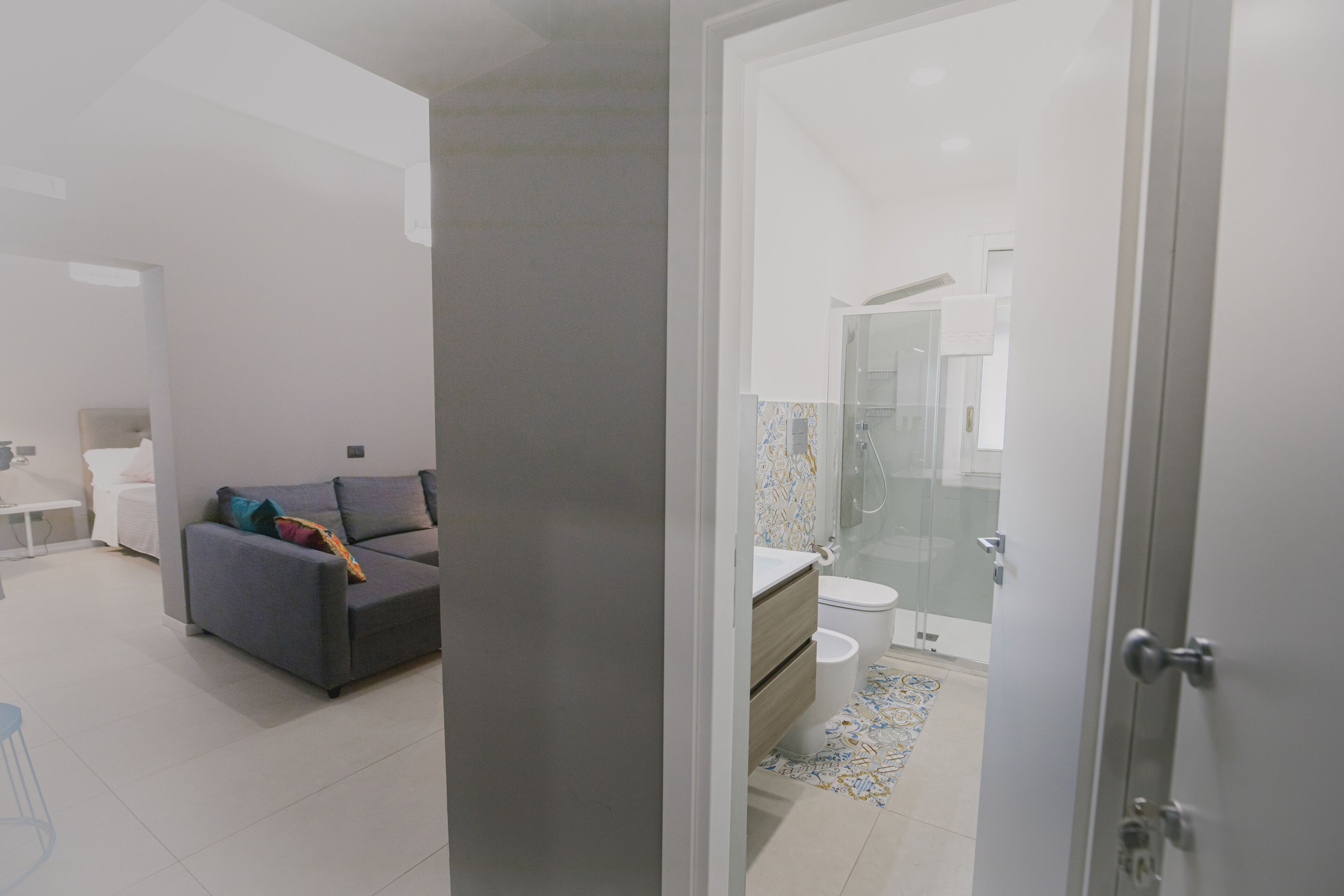 A modern hotel room with a grey sofa, a small table, and a bathroom with a glass shower enclosure.